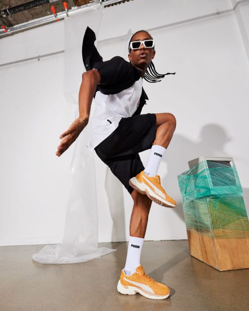 Person in mid-air wearing Puma athletic shoes, black shorts, and white sunglasses, with retro-style computer in the background.