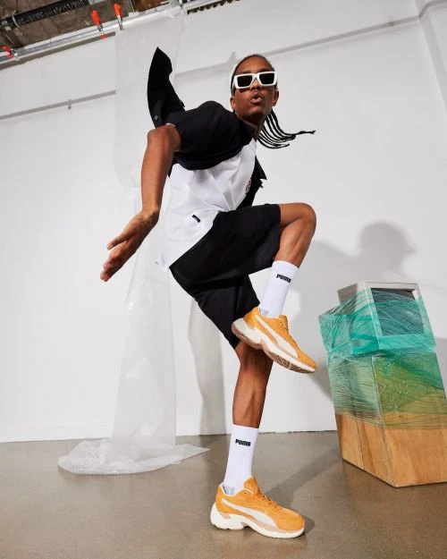 Person in mid-air wearing Puma athletic shoes, black shorts, and white sunglasses, with retro-style computer in the background.