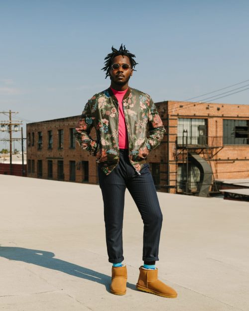 A male model with dreadlocks and sunglasses poses on a rooftop wearing a floral jacket, pink shirt, navy pants, and Ugg boots.