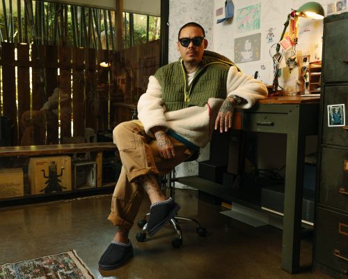 Man with tattoos in olive and cream jacket poses in slippers, sitting in front of desk in a brightly lit room with bamboo and art.