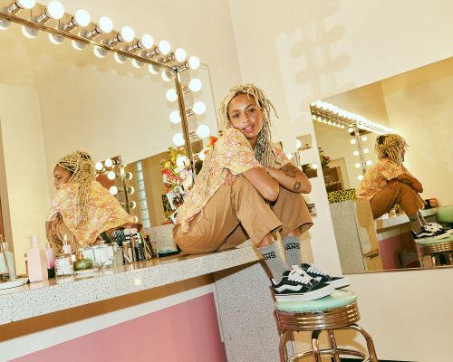 Stylish woman with blonde dreadlocks poses in a beauty room, wearing a floral top and Vans sneakers, sitting on a counter near a lighted mirror.