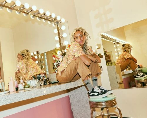 Stylish woman with blonde dreadlocks poses in a beauty room, wearing a floral top and Vans sneakers, sitting on a counter near a lighted mirror.