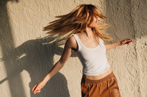 Woman with long blonde hair tossing her head against a textured beige stucco wall, wearing a white tank top and brown skirt.