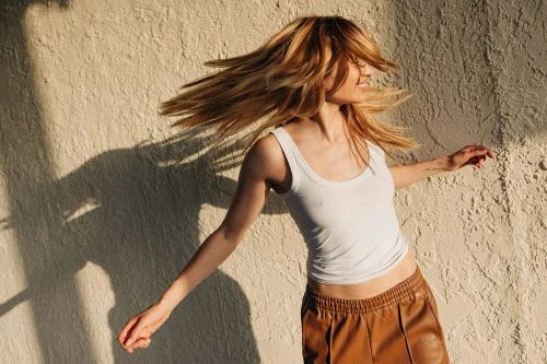 Woman with long blonde hair tossing her head against a textured beige stucco wall, wearing a white tank top and brown skirt.