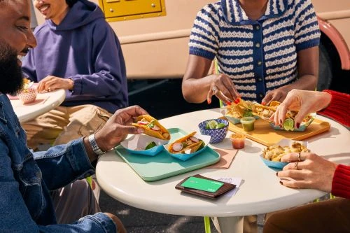 Group of people eating tacos at a white table outside a food truck, enjoying a casual dining experience with vibrant colors and fresh ingredients.