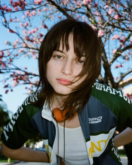 A young adult female poses in front of a tree with pink flowers. She wears a zip-up jacket and has headphones around her neck.