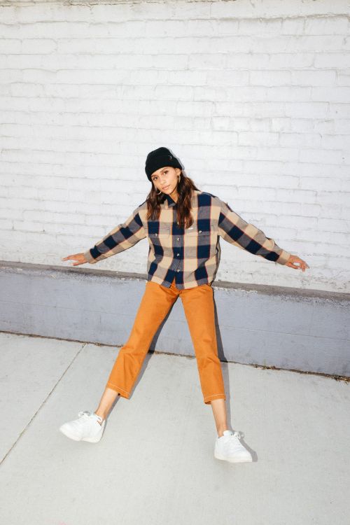 Full-length shot of a young adult woman outdoors wearing a flannel shirt, orange pants, a beanie, and sneakers posing with outstretched arms.