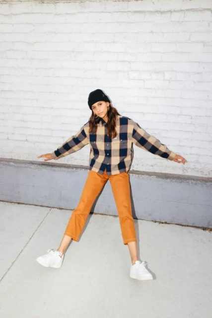 Full-length shot of a young adult woman outdoors wearing a flannel shirt, orange pants, a beanie, and sneakers posing with outstretched arms.