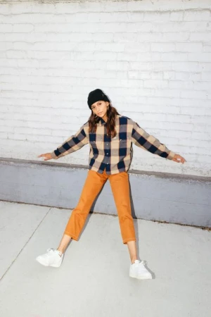 Full-length shot of a young adult woman outdoors wearing a flannel shirt, orange pants, a beanie, and sneakers posing with outstretched arms.
