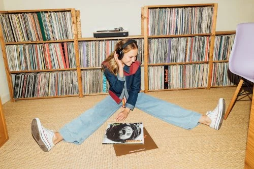 Young woman with headphones sits with legs spread in front of a record collection, listening to vinyl in a relaxed indoor setting.