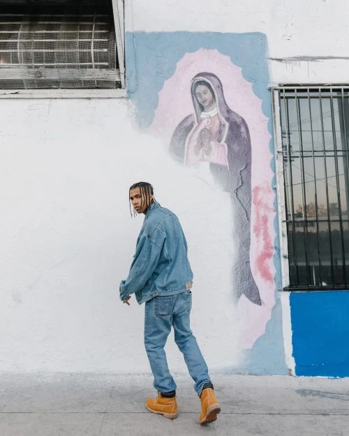 Young man in blue denim jacket and jeans walks past a vibrant wall mural in an urban setting, showcasing contemporary street style.