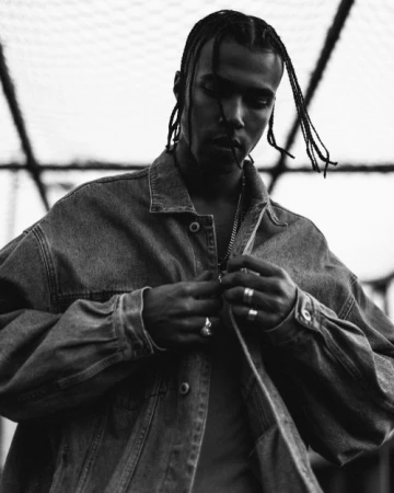 Monochrome portrait of a Black man with braided hair wearing a denim jacket and rings, photographed by Neil Favila