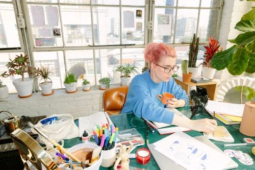 Woman with pink hair in blue sweater drinks from an orange mug while drawing at a desk covered in art supplies near a window.