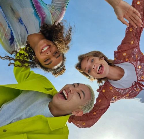Low-angle shot of three smiling young adults looking down toward the camera against a bright blue sky. Diverse group with a positive mood.