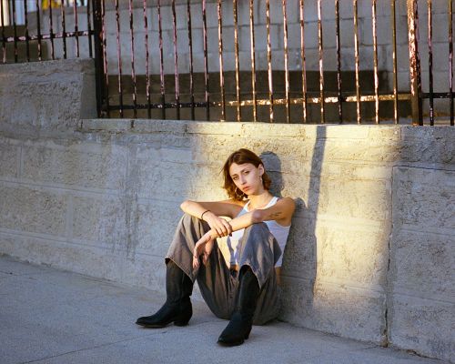A young woman with short brown hair sitting on the ground, leaning against a stone wall with a wrought iron fence above.