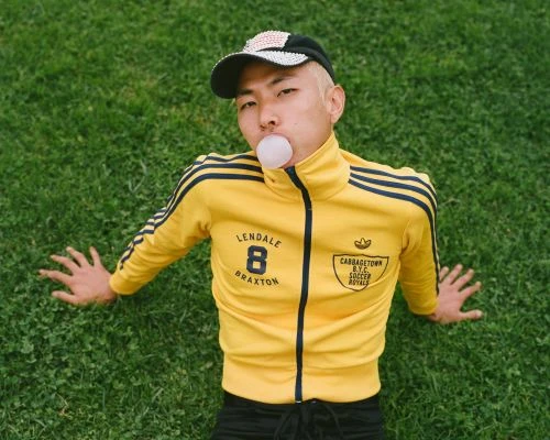 Person blowing bubblegum bubble lying on grass wearing yellow jacket and baseball cap with vintage vibes and Adidas logo.