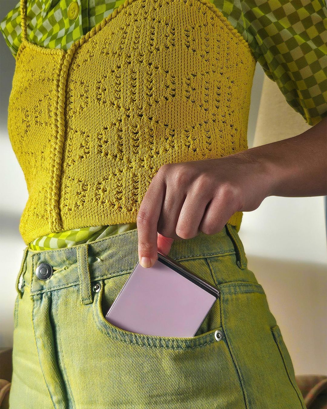 Woman puts a pink smartphone into her green-yellow jeans pocket. She wears a yellow knit top and a green patterned top.