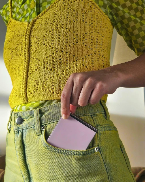 Woman puts a pink smartphone into her green-yellow jeans pocket. She wears a yellow knit top and a green patterned top.
