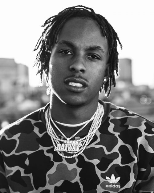 Black and white portrait of a young Black man with dreadlocks wearing a patterned sweatshirt and layered diamond chain necklaces.