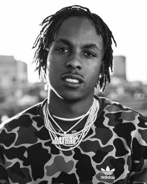 Black and white portrait of a young Black man with dreadlocks wearing a patterned sweatshirt and layered diamond chain necklaces.