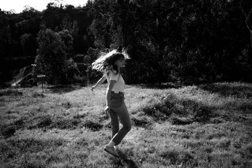 Monochrome image of a woman with long hair spinning on a grassy hill surrounded by trees, captured by Neil Favila.