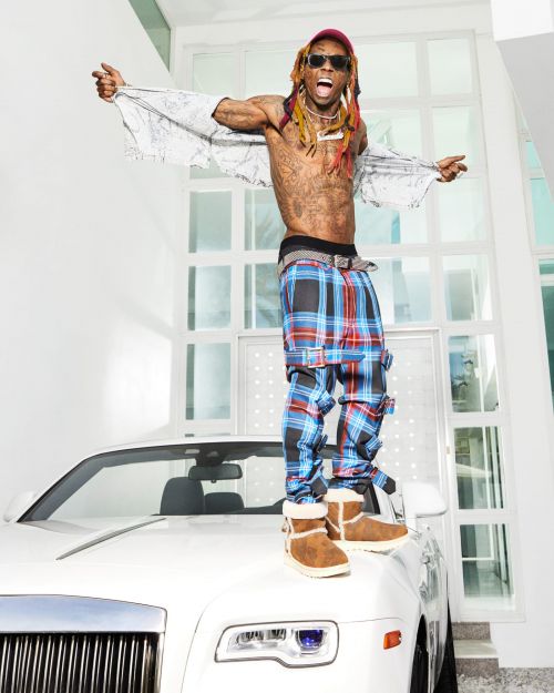 Lil Wayne strikes a pose on a white car, wearing plaid pants, brown boots, and a baseball cap, against a white building background.