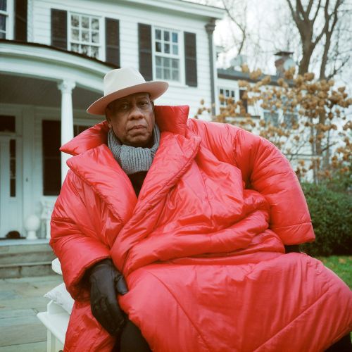Adult male wrapped in a large red coat sitting outside, wearing a hat and scarf, with a white house and trees in the background.