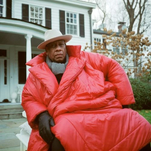 Adult male wrapped in a large red coat sitting outside, wearing a hat and scarf, with a white house and trees in the background.