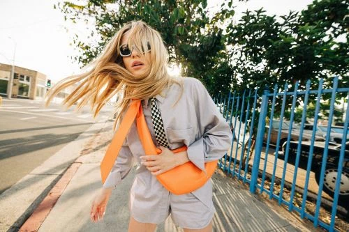 A stylish woman with blonde hair blowing in the wind stands on a city sidewalk wearing sunglasses, shorts, a shirt, and an orange purse.