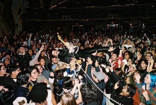 A musician crowd surfing with his guitar during a concert, supported by a large, cheering audience in a dimly lit music venue.