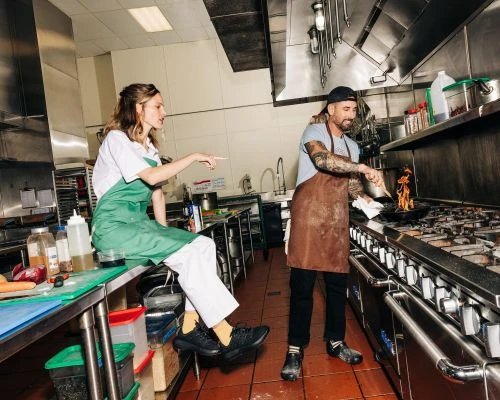 Two people, a man and a woman, in a commercial kitchen. The man cooks with flames while the woman sits and points with an apron.