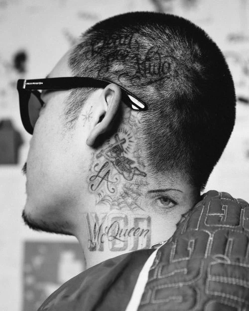 Close-up black and white portrait of an adult male with dark hair, wearing dark glasses and featuring numerous tattoos on his head and neck.