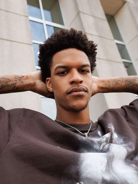Portrait of an adult male with dark curly hair and tattoos, wearing a brown sweatshirt and silver necklace, posing outdoors against an office building.