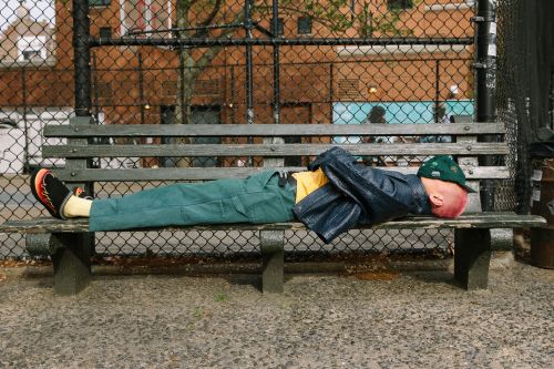 Adult lies prone on a park bench outdoors, wearing a navy jacket, green pants, baseball cap, and colourful socks, with a chainlink fence behind.