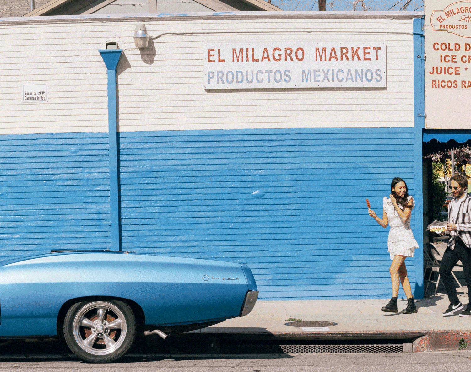 Man and woman walking past a blue and white painted market wall with a vintage blue car parked in the foreground