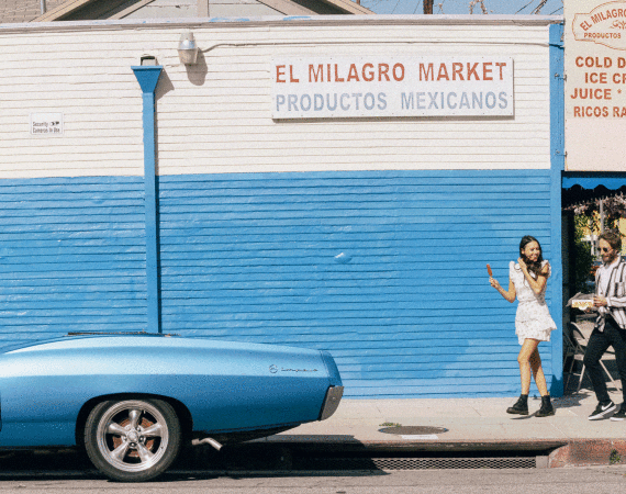 Man and woman walking past a blue and white painted market wall with a vintage blue car parked in the foreground