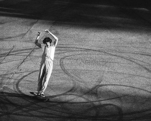 Monochrome action shot of skateboarder in overalls with arms raised, riding on asphalt with tire marks, creating a dynamic black and white image.