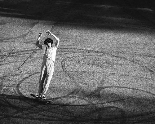 Monochrome action shot of skateboarder in overalls with arms raised, riding on asphalt with tire marks, creating a dynamic black and white image.