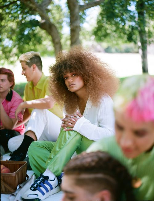 Outdoor picnic with a group of young adults, including a woman with afro hair wearing green pants and sneakers in a grassy park.
