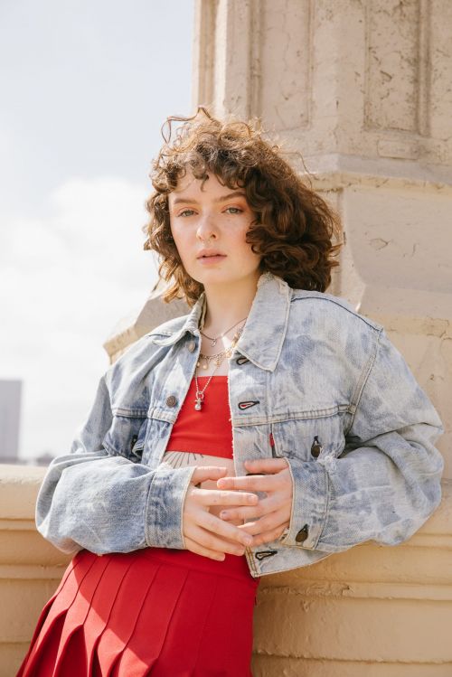 Young woman with curly hair wearing a jean jacket and red skirt, posing next to a stone pillar on a sunny day in an outdoor setting.