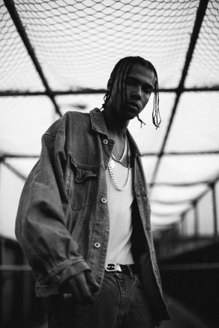 Monochrome portrait of a young adult with braided hair wearing denim jacket and silver chains against a geometric structure backdrop.