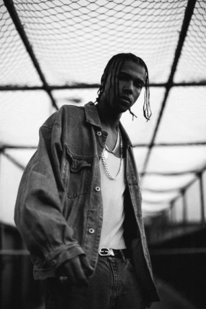 Monochrome portrait of a young adult with braided hair wearing denim jacket and silver chains against a geometric structure backdrop.