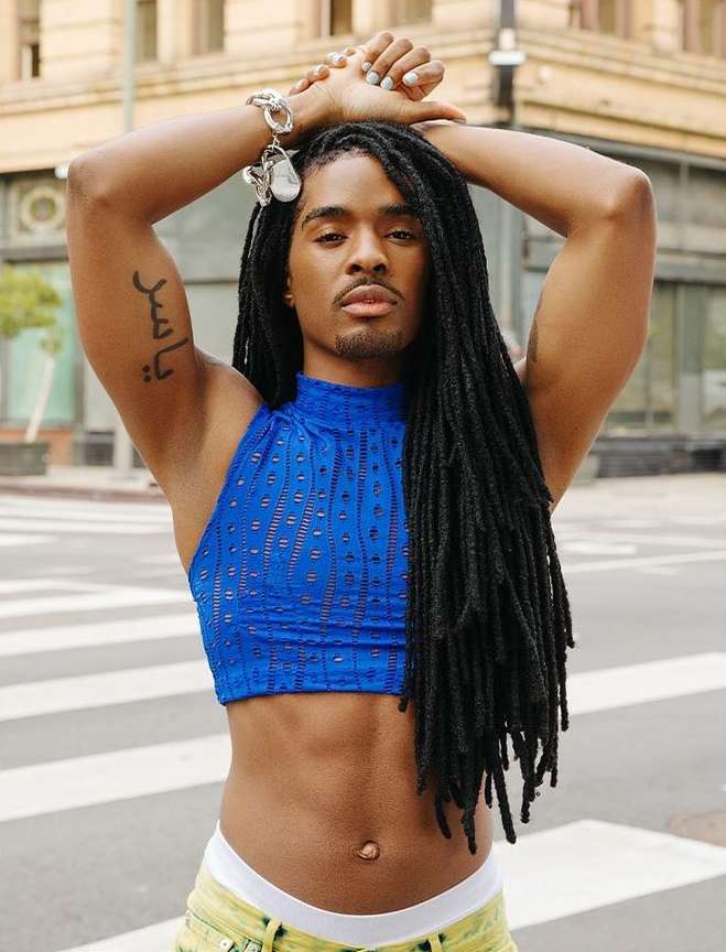 An adult male with long dreadlocks poses in a blue top and yellow pants on a city street, showcasing fashion and personal style.