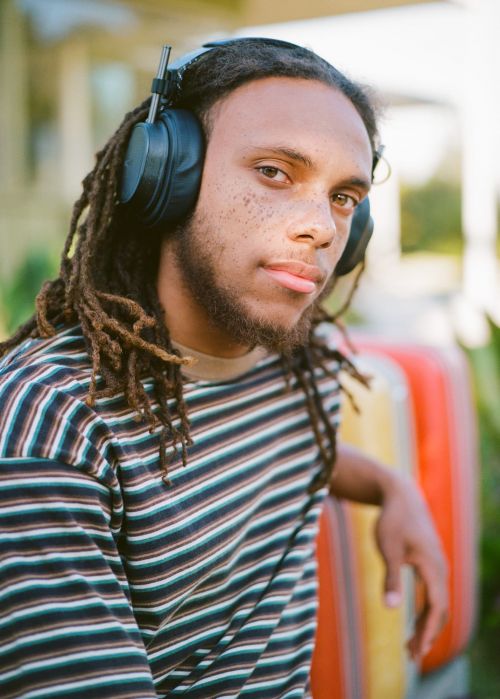 A young man with dreadlocks and headphones wearing a striped shirt outdoors, with a relaxed expression and natural lighting.