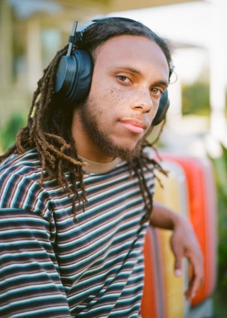 A young man with dreadlocks and headphones wearing a striped shirt outdoors, with a relaxed expression and natural lighting.