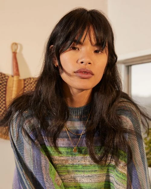 Portrait of a young adult woman with dark hair and bangs wearing a colorful striped sweater indoors, looking at the camera.