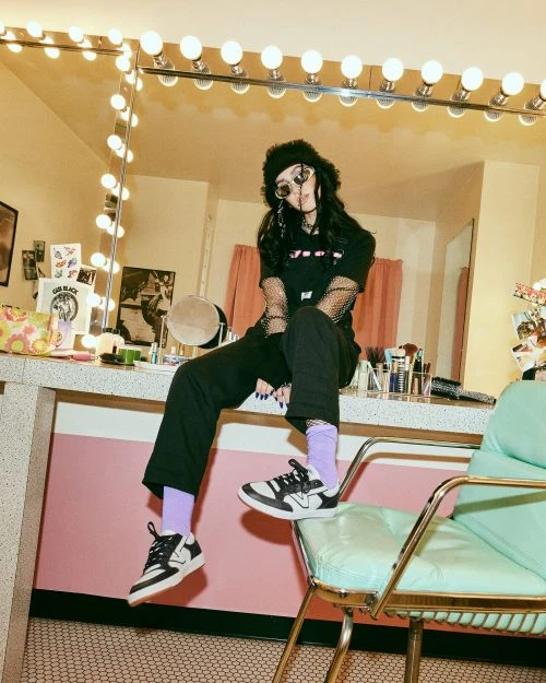 Young woman in black streetwear sits on a vintage chair in a retro dressing room with a large mirror and bulb lighting.