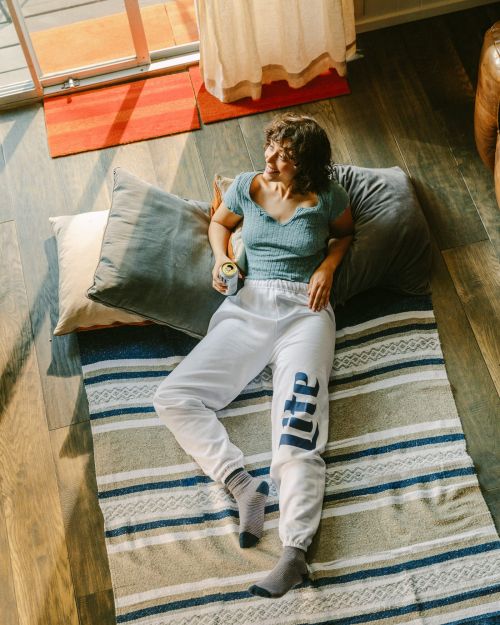 Young woman in sweatpants lying on a striped blanket with cushions, holding a beer can and looking out the window in indoor daylight.