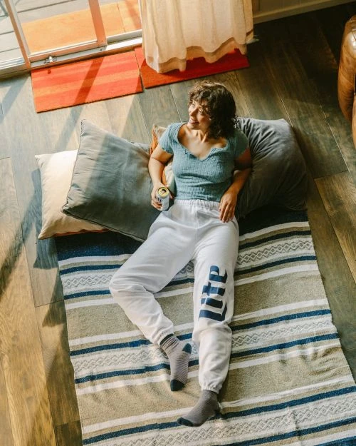 Young woman in sweatpants lying on a striped blanket with cushions, holding a beer can and looking out the window in indoor daylight.