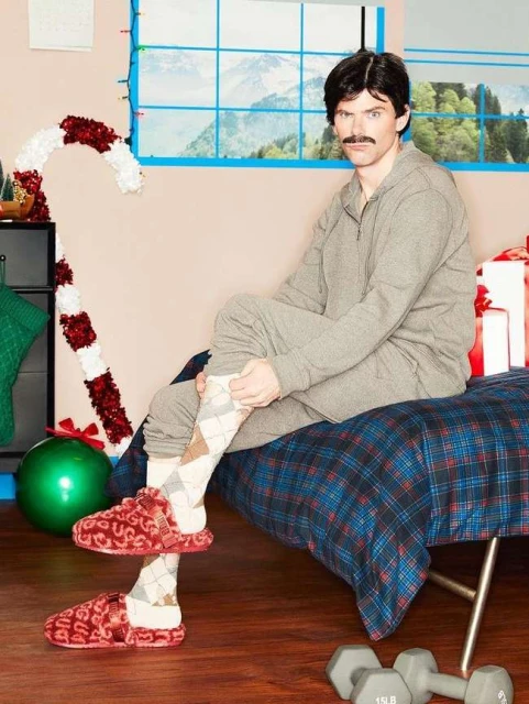 Man in grey sweatsuit sitting on bench near Christmas decorations, wearing argyle socks and fluffy slippers. Dumbbells visible on the floor.